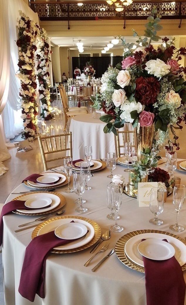 A round table at a wedding reception features gold chargers, white plates, deep red napkins, and a floral centerpiece.