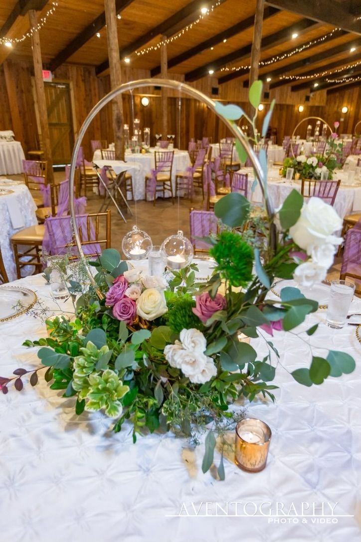 A floral centerpiece with a gold hoop and white and purple roses sits on a table in a rustic banquet hall with fairy lights.
