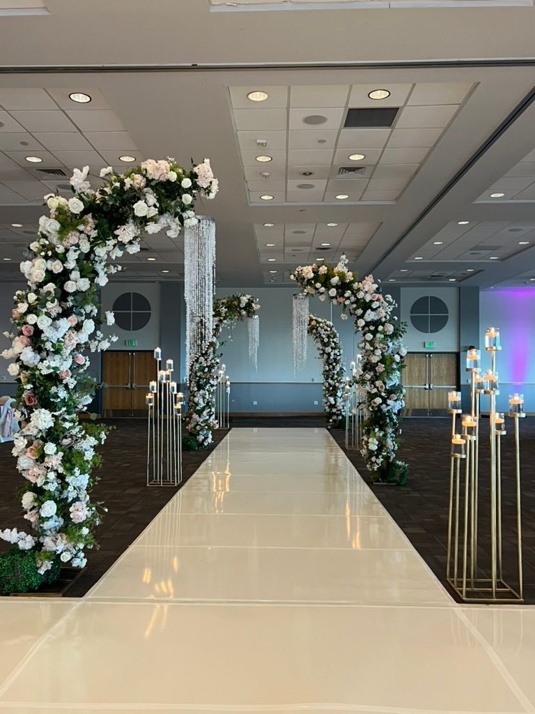 A glossy white aisle lined with tall gold candle stands and large floral arches decorated with white and pink flowers.