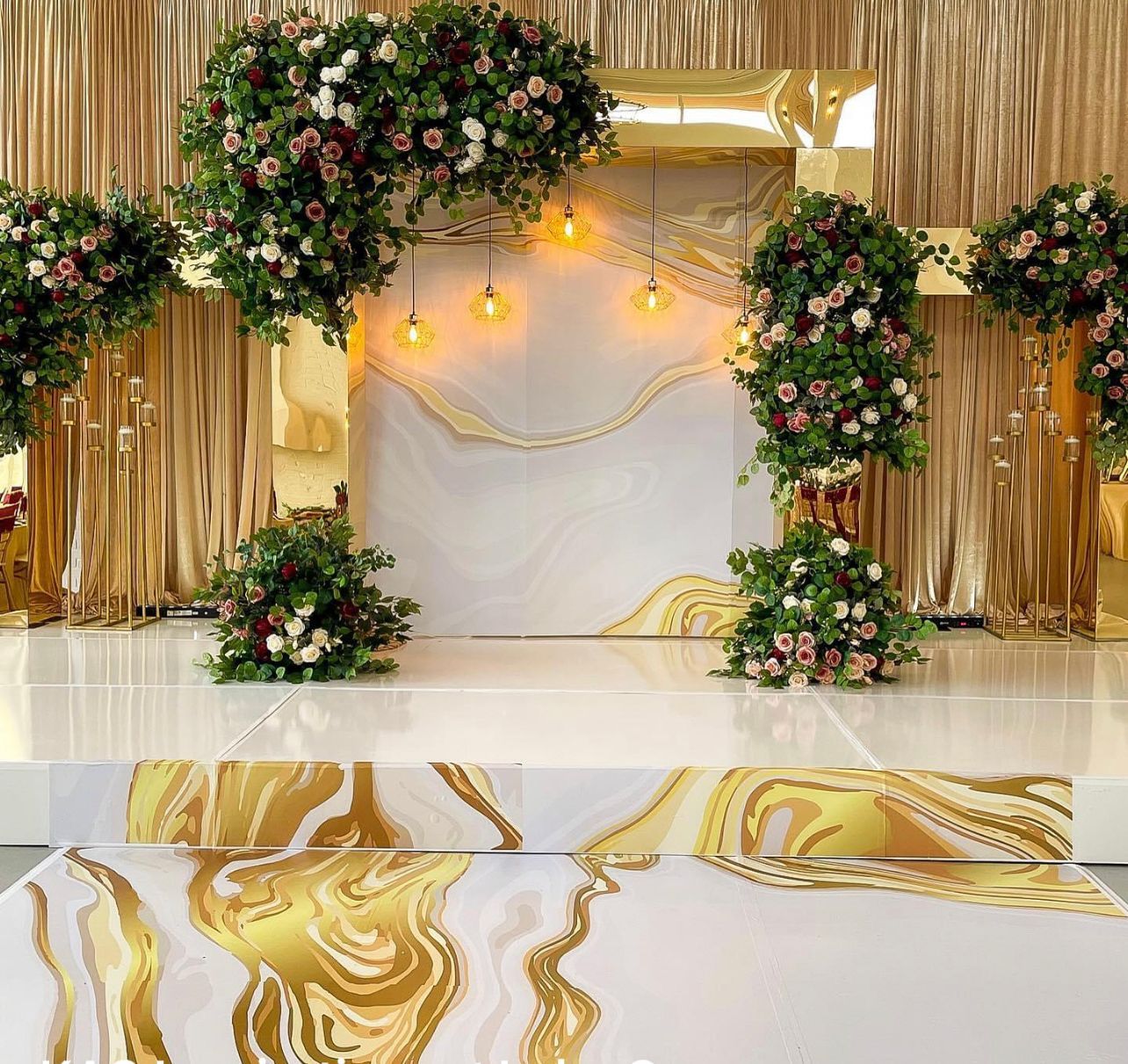 A wedding stage featuring gold fringe curtains, marble-patterned decor, floral arrangements, and hanging Edison bulbs.