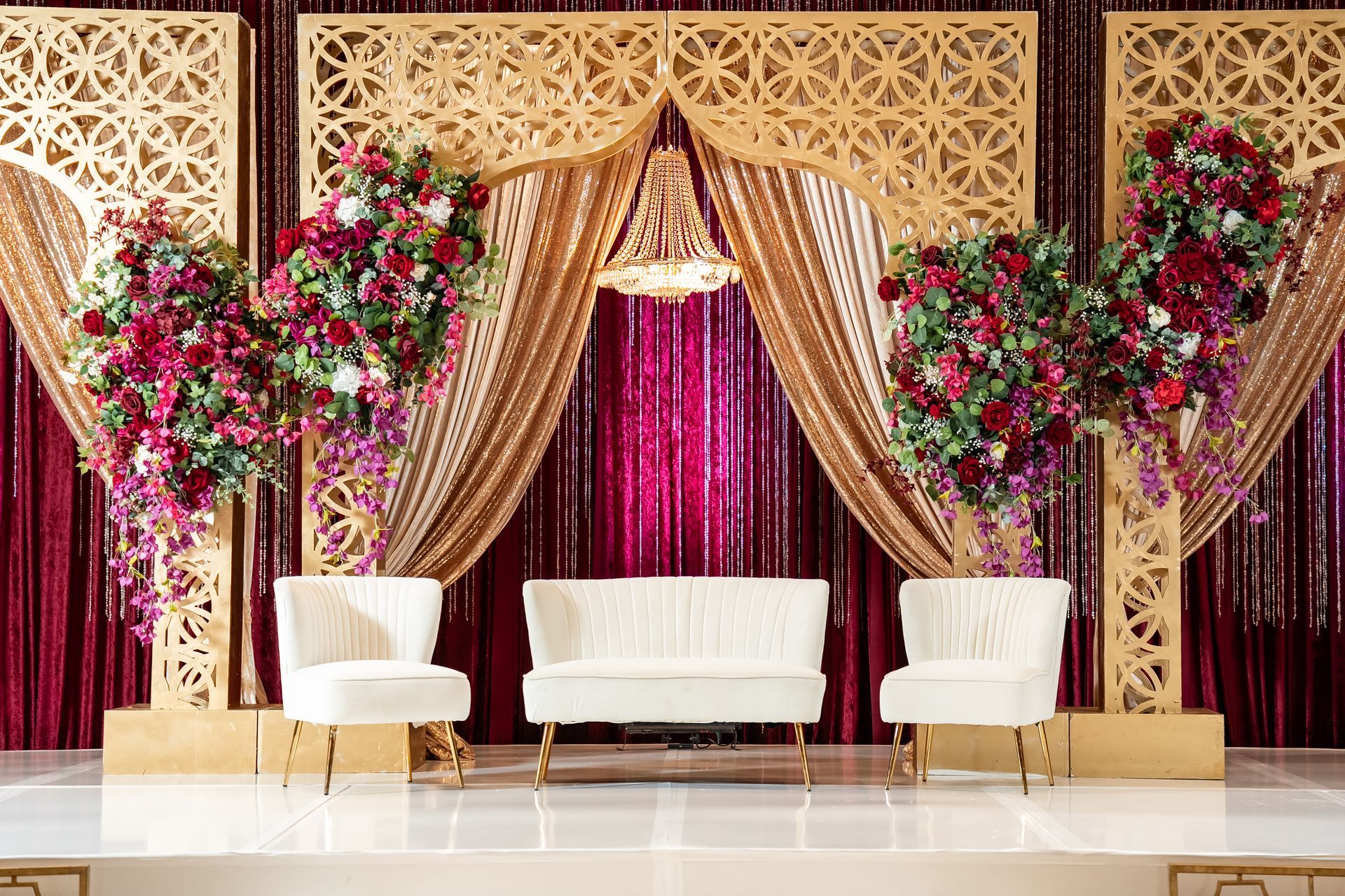 Elegant stage setup with a white sofa and two chairs, framed by gold lattice screens, floral arrangements, and red drapes.