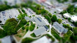 Aerial view of a building surrounded by trees, parking lot, and road with blurred cars. Green and white color scheme.