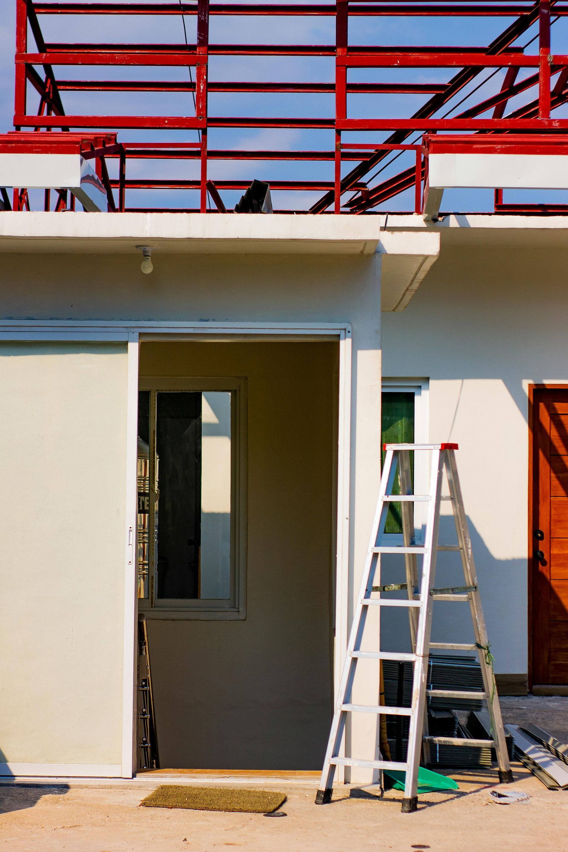 A house under construction; a white wall, a window and a ladder in front of the open doorway. Red metal frame roof.