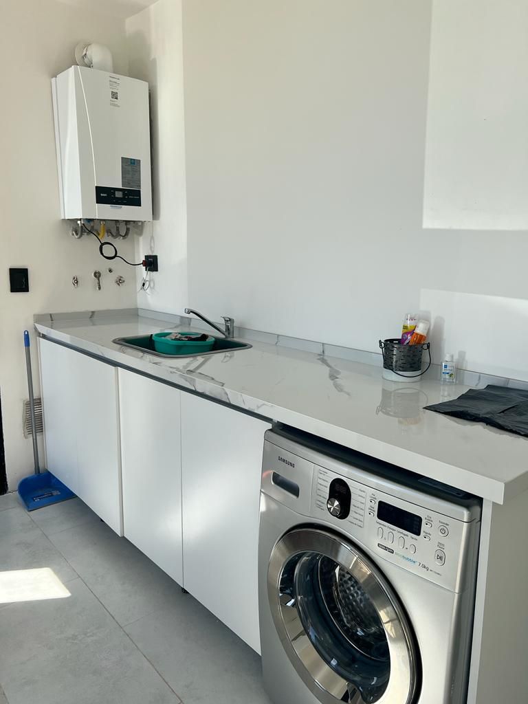 Cuarto de lavandería con armarios blancos, lavadora, fregadero y calentador de agua en la pared.