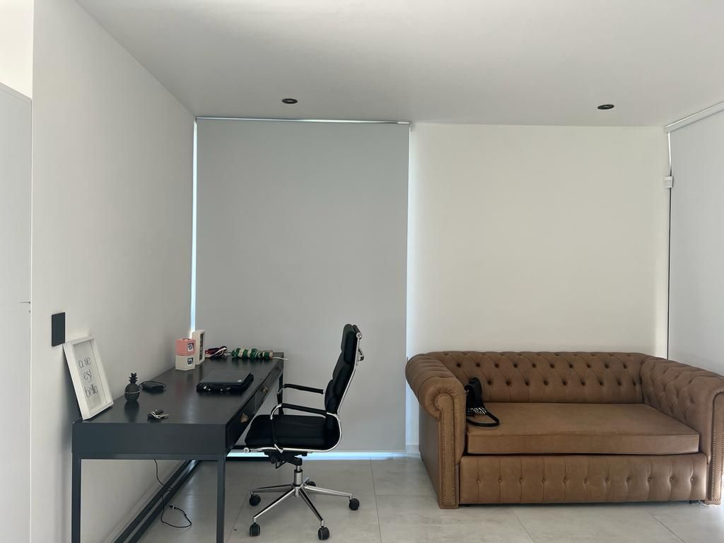 Espacio de oficina con escritorio negro, silla y sofá color beige. Paredes blancas y persianas.