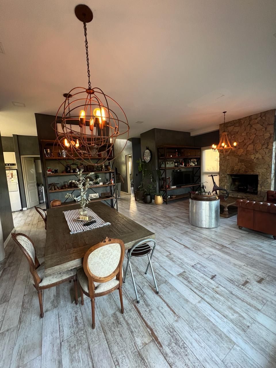 Espacio abierto que integra comedor y sala de estar, con mesa rústica de madera, lámparas ornamentadas y chimenea de piedra.