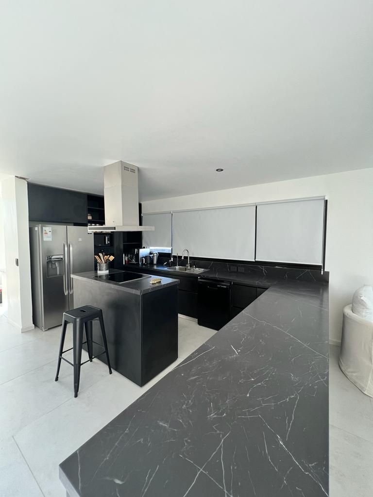 Cocina moderna con encimeras negras, electrodomésticos de acero inoxidable e isla. Paredes blancas, grandes ventanales.