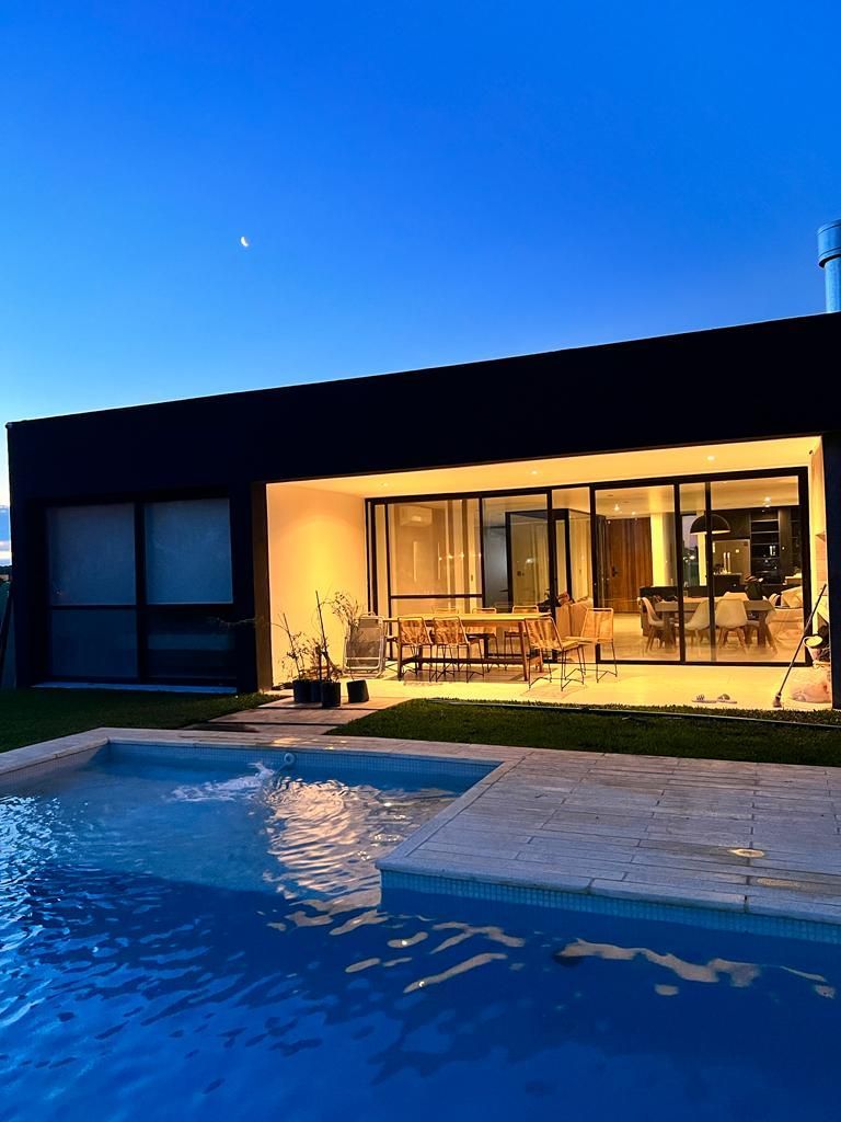 Piscina y casa moderna al atardecer; interior iluminado visible a través de puertas corredizas de vidrio.