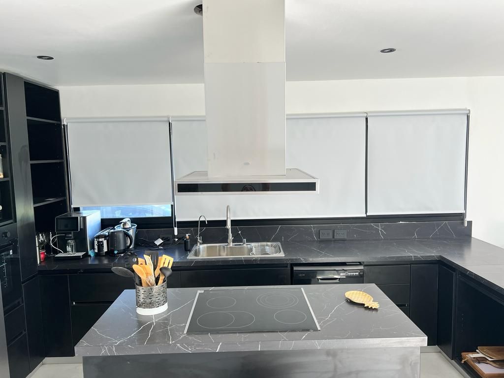Cocina moderna con armarios y encimeras negras, isla con placa de cocina, campana extractora blanca y persianas enrollables.