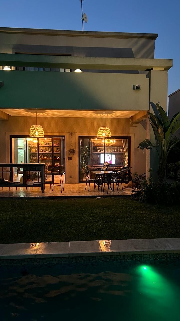 Vista del patio trasero de una casa al atardecer, con piscina, patio e interior iluminado visibles.
