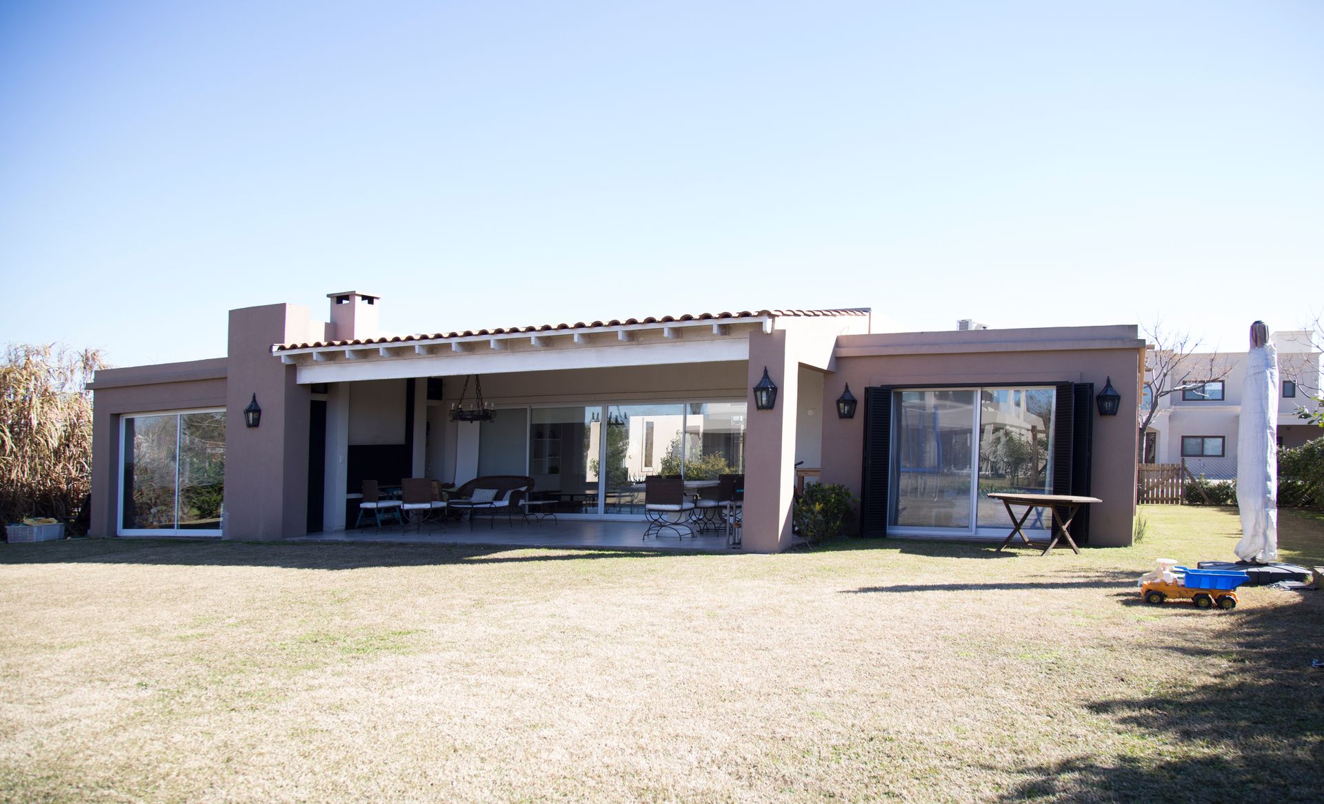 Una casa baja, de color marrón, con grandes ventanas y un patio abierto sobre un césped bajo un cielo despejado.