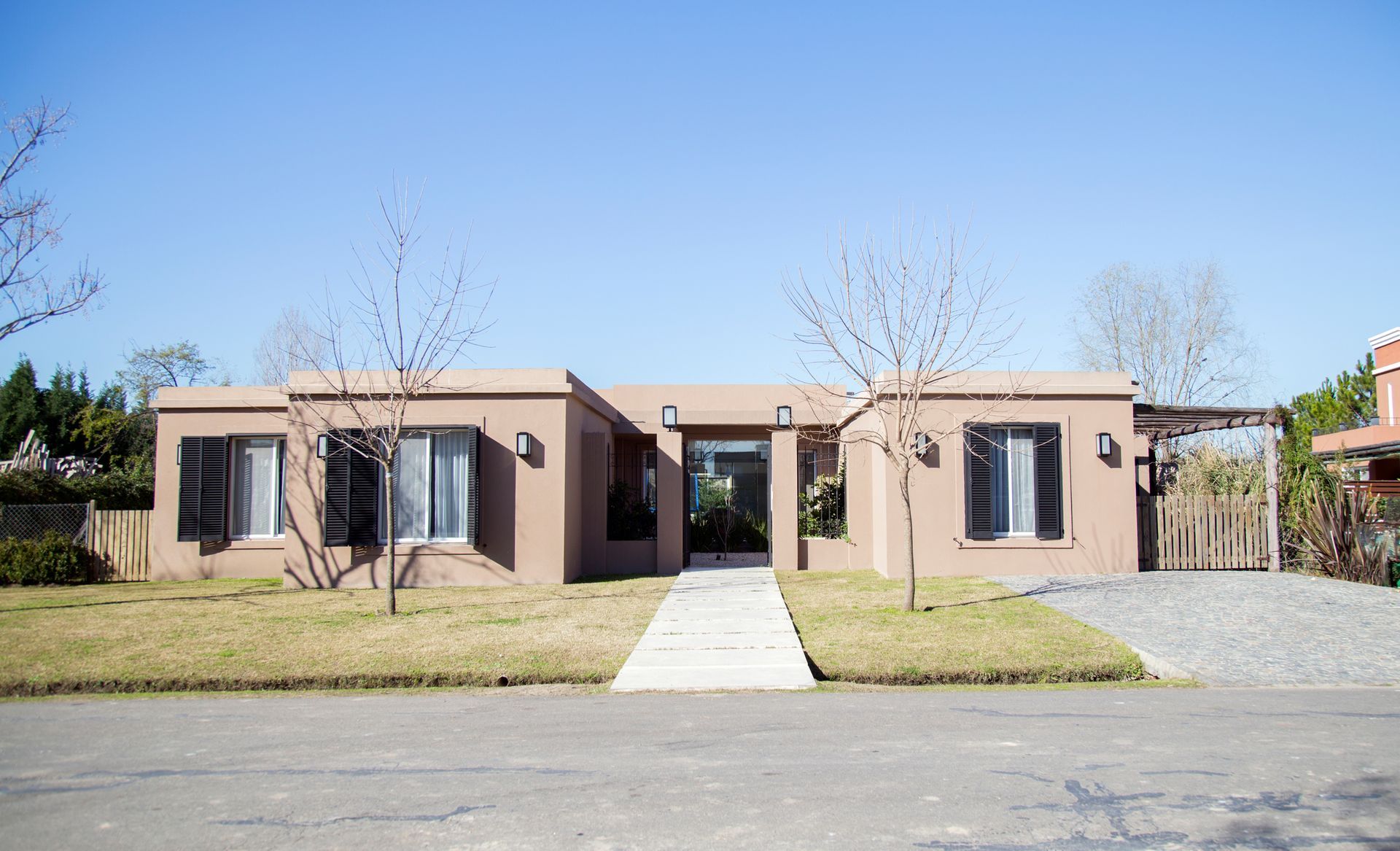Casa de estuco marrón con una acera de hormigón y dos pequeños árboles desnudos en un día soleado.