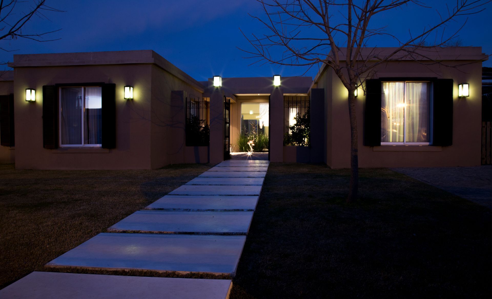 Casa color beige de noche, iluminada por luces exteriores; un sendero conduce a la puerta principal.