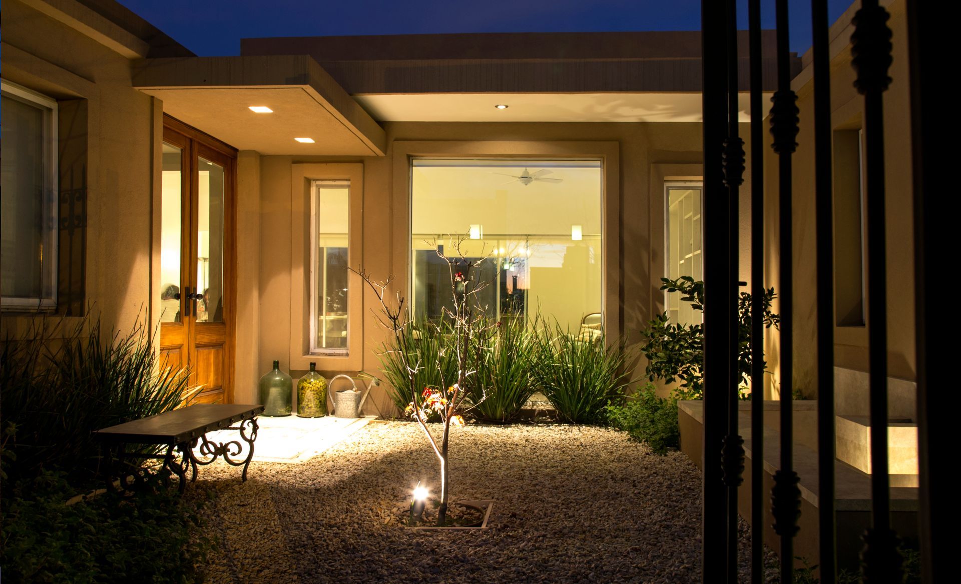 Patio de entrada iluminado por la noche, edificio color beige, grava, banco