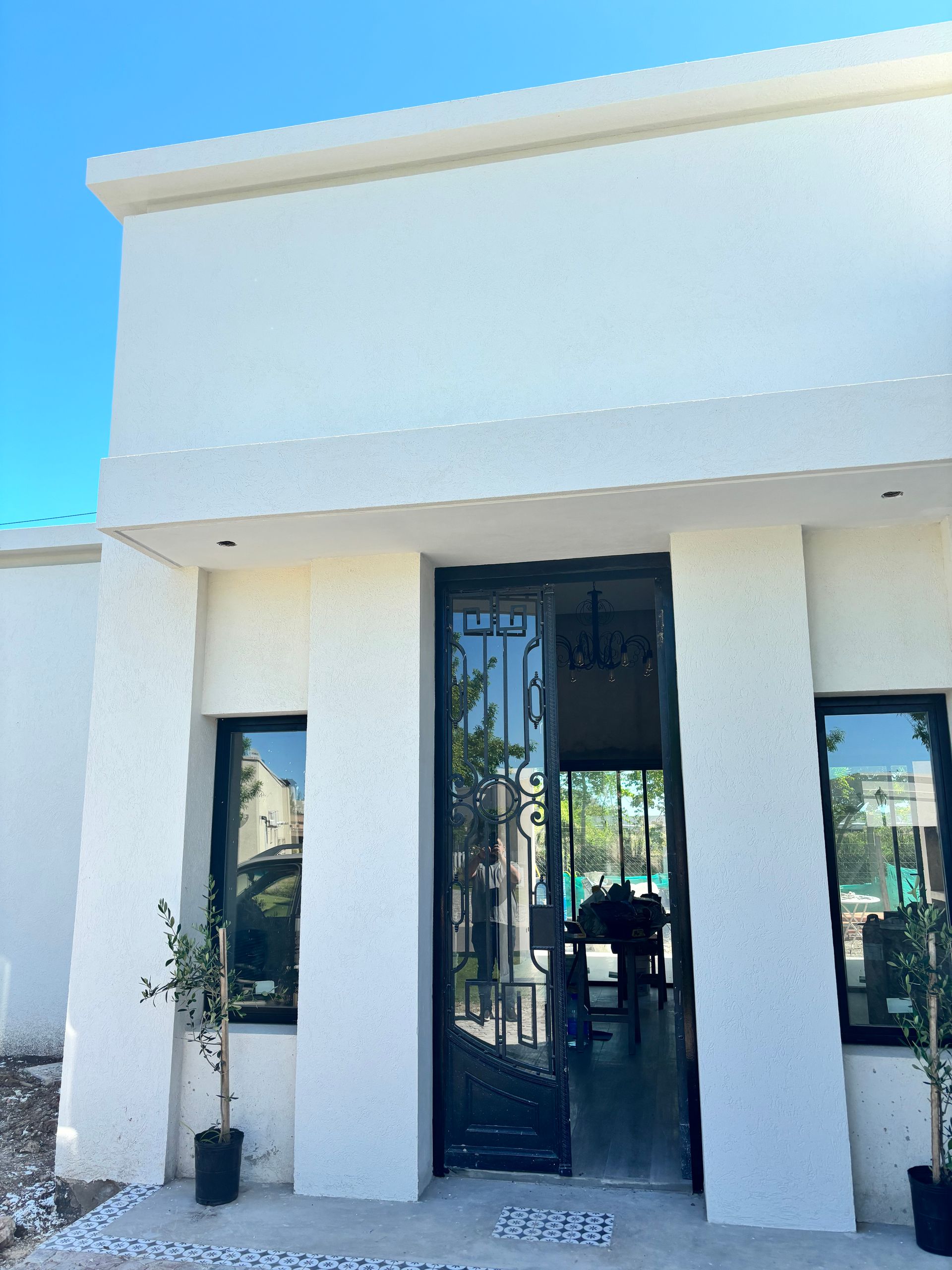 Entrada de edificio blanco con puerta y ventanas negras, plantas en macetas y cielo azul despejado.