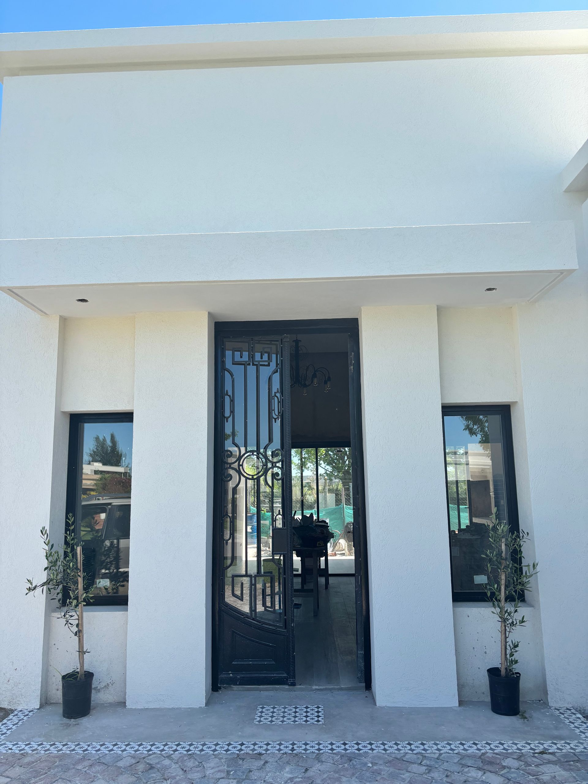 Edificio blanco con puerta y ventanas negras; dos pequeños árboles flanquean la entrada.