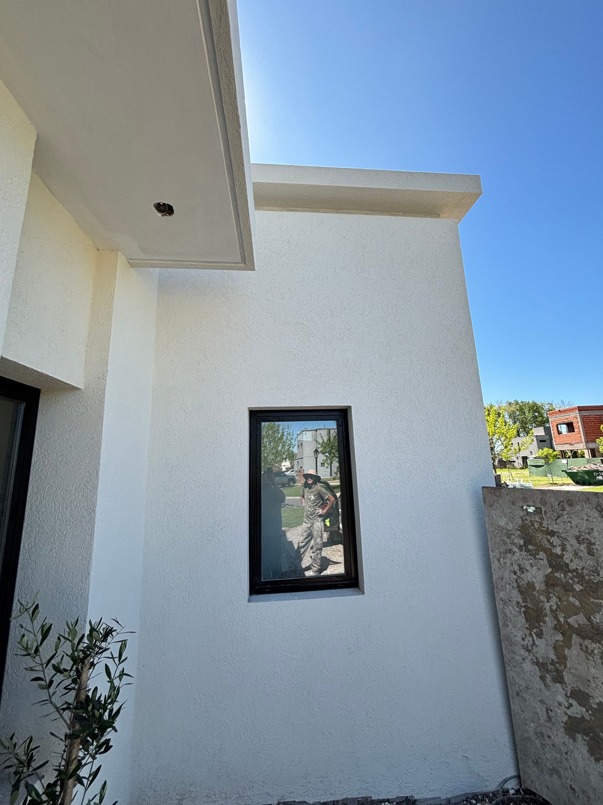 Exterior de edificio blanco con ventana de marco negro que refleja los árboles, bajo un cielo azul.