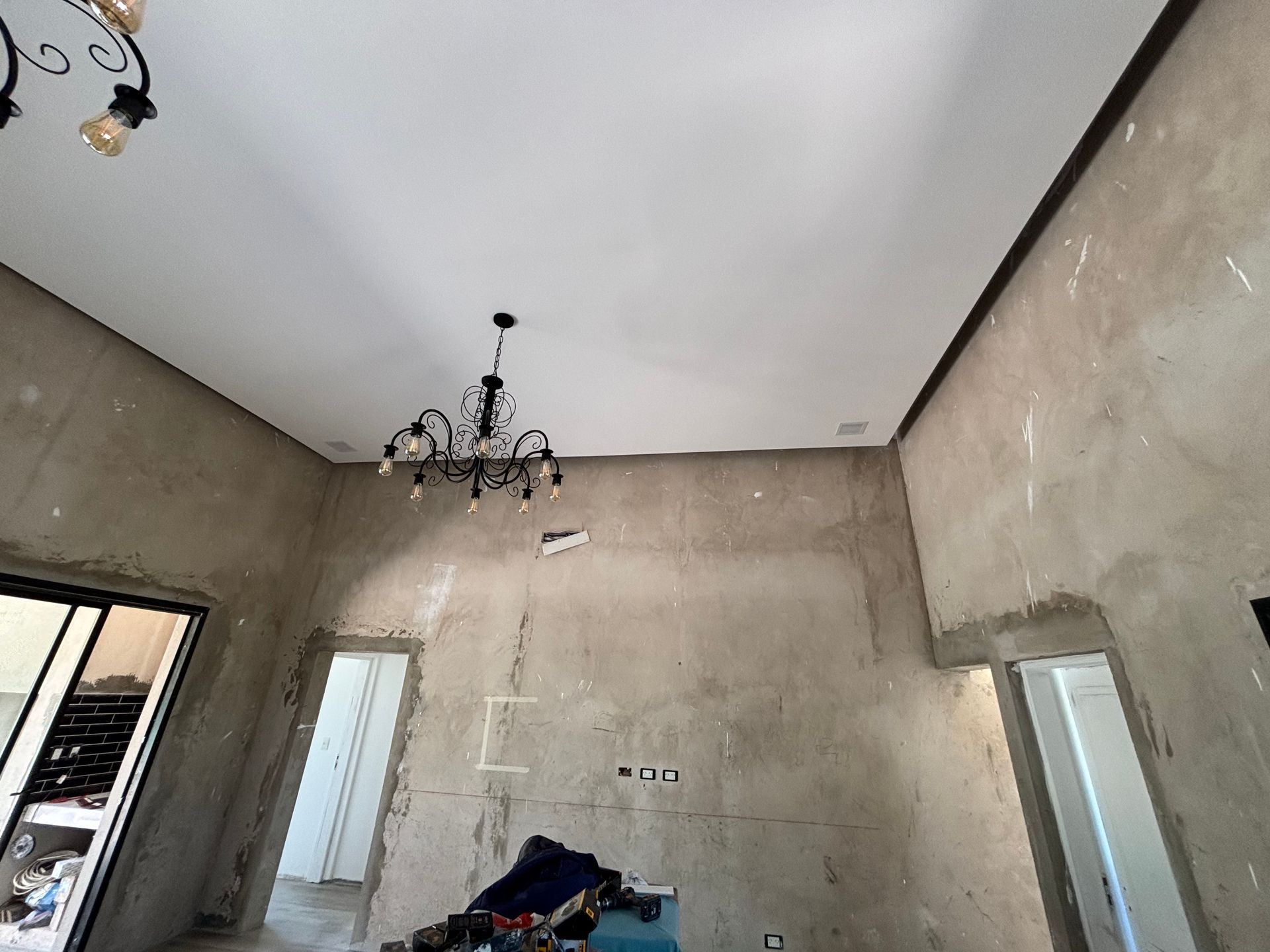 Habitación en construcción con paredes de hormigón visto, techo blanco y lámpara de araña.