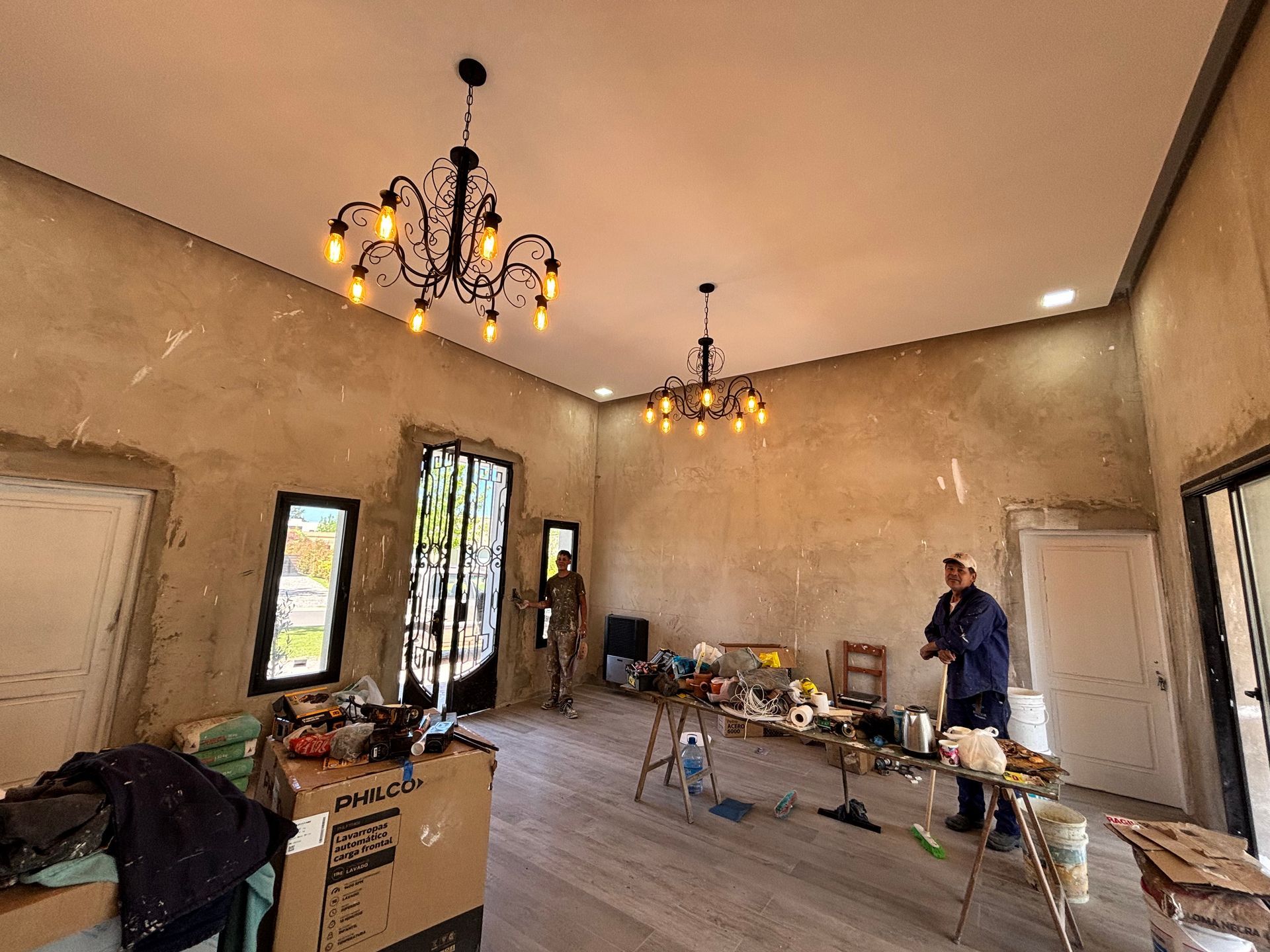 Habitación con dos candelabros, paredes sin terminar y una persona de pie cerca de una mesa con varios objetos.