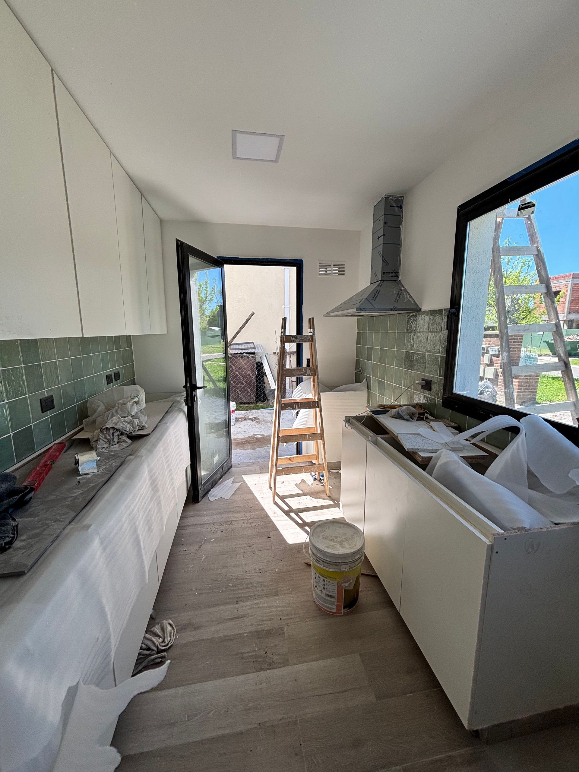 Cocina en construcción con armarios blancos, puerta abierta, escalera y campana extractora.