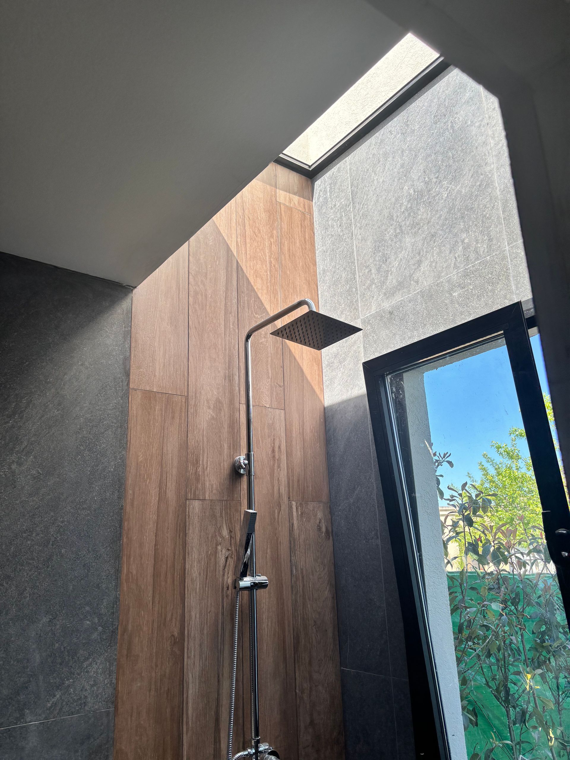 Ducha con paneles de madera y tragaluz. Cabezal de ducha de acero inoxidable, ventana con vistas a la vegetación.