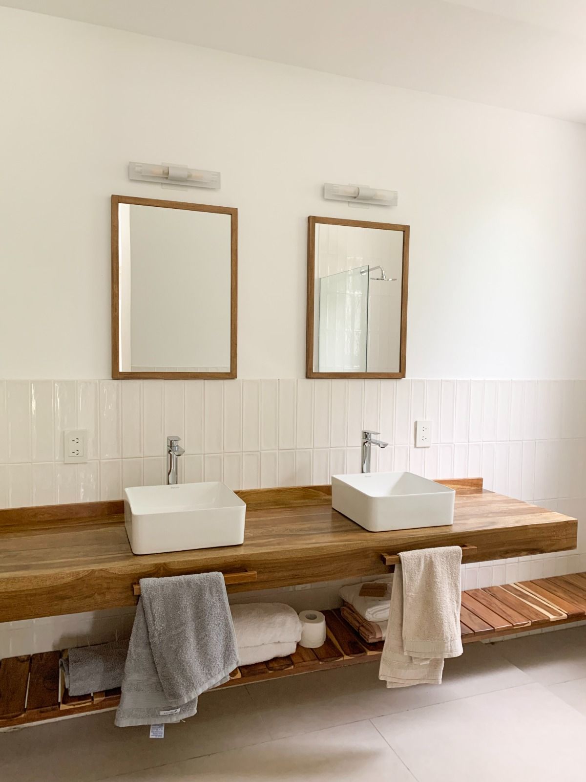 Baño con dos lavabos cuadrados blancos sobre una encimera de madera, espejos enmarcados y toalleros.