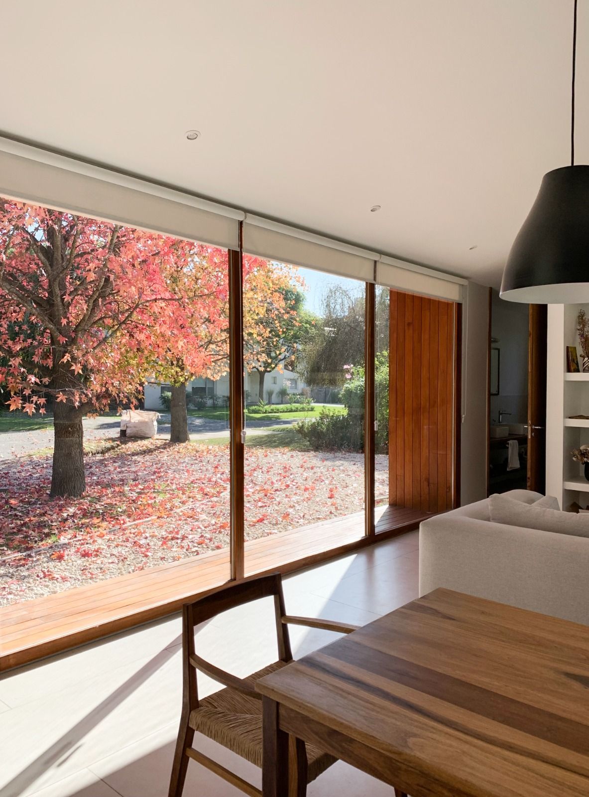 Comedor con mesa de madera y gran ventanal con vistas a árboles otoñales con hojas rojas.