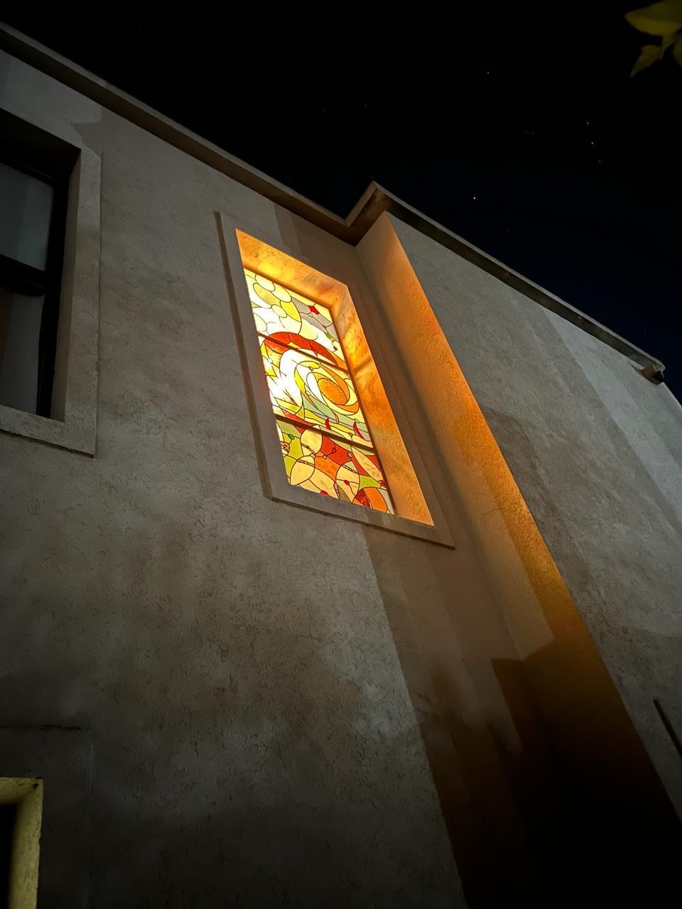Exterior del edificio de noche con una vidriera iluminada; una luz cálida ilumina la pared.