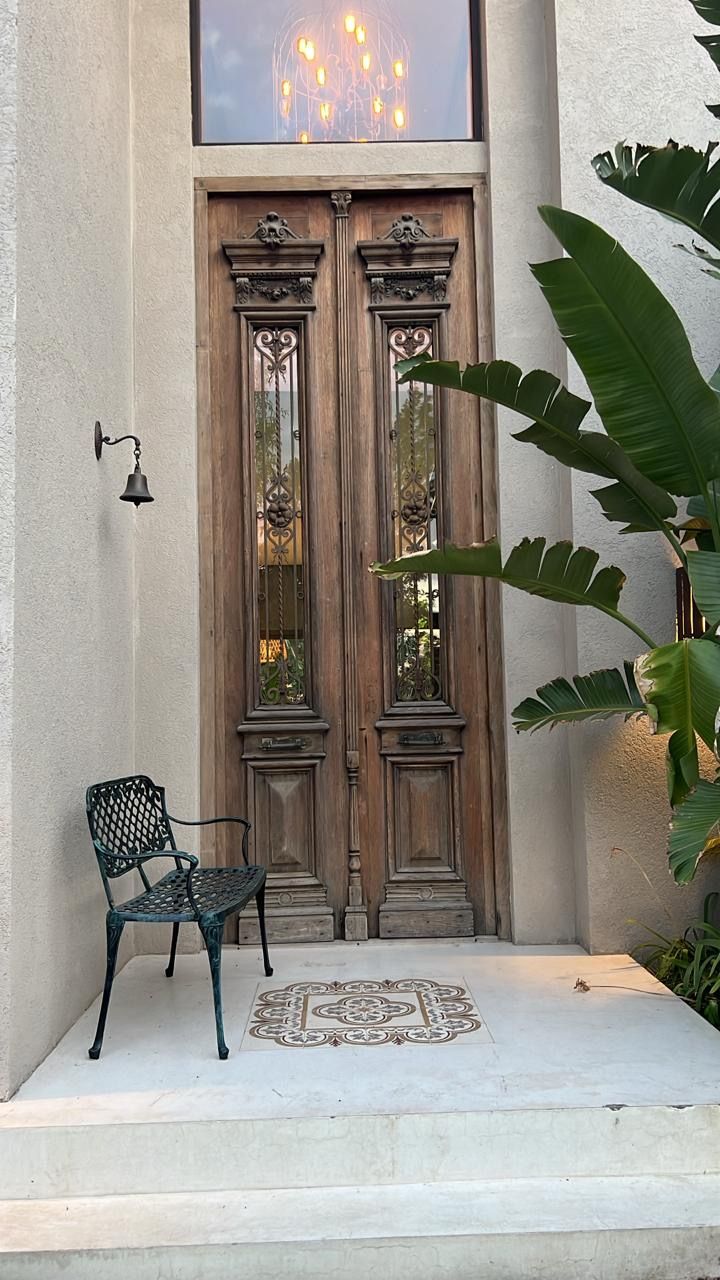 Puertas dobles de madera con intrincadas tallas y paneles de vidrio, flanqueadas por plantas y una silla de hierro forjado.