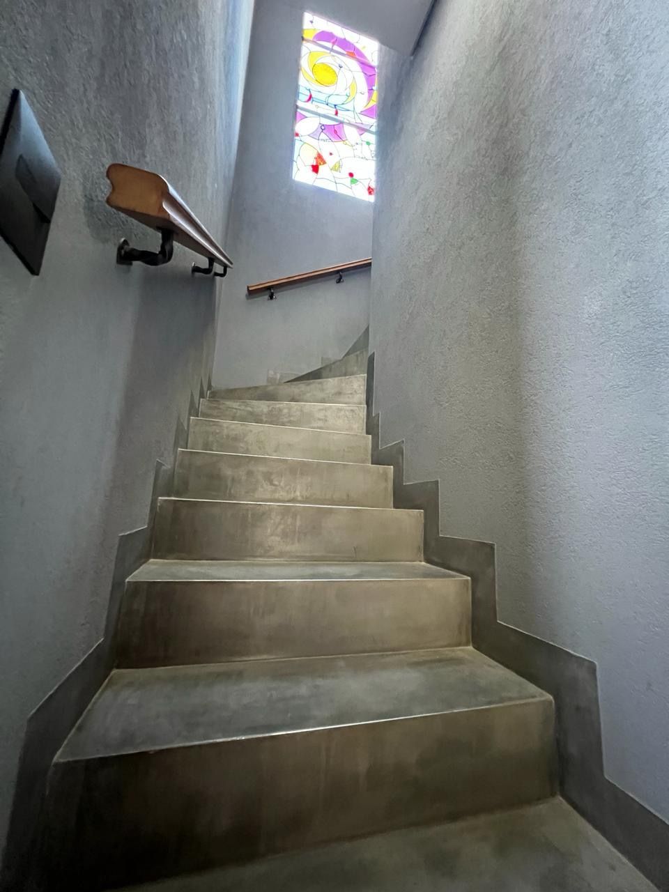 Escalera de hormigón que asciende, con una vidriera en la parte superior. Paredes grises y barandilla de madera.