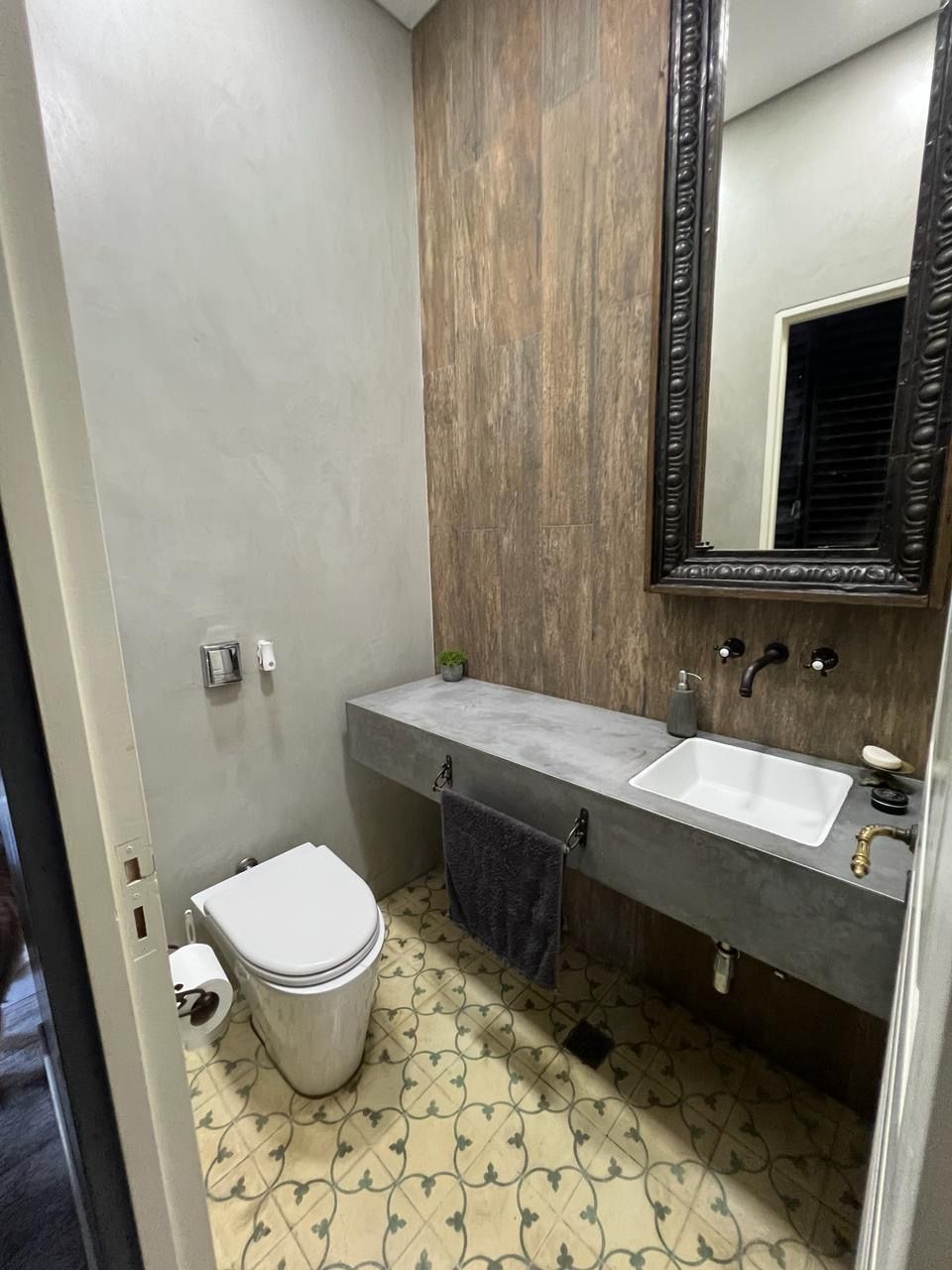 Tocador con detalles en gris y madera, lavabo de hormigón, suelo de baldosas estampadas y espejo ornamentado.