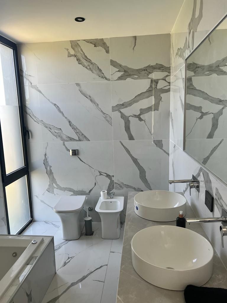 Baño moderno con azulejos imitación mármol, que incluye inodoro, bidé, dos lavabos y bañera.
