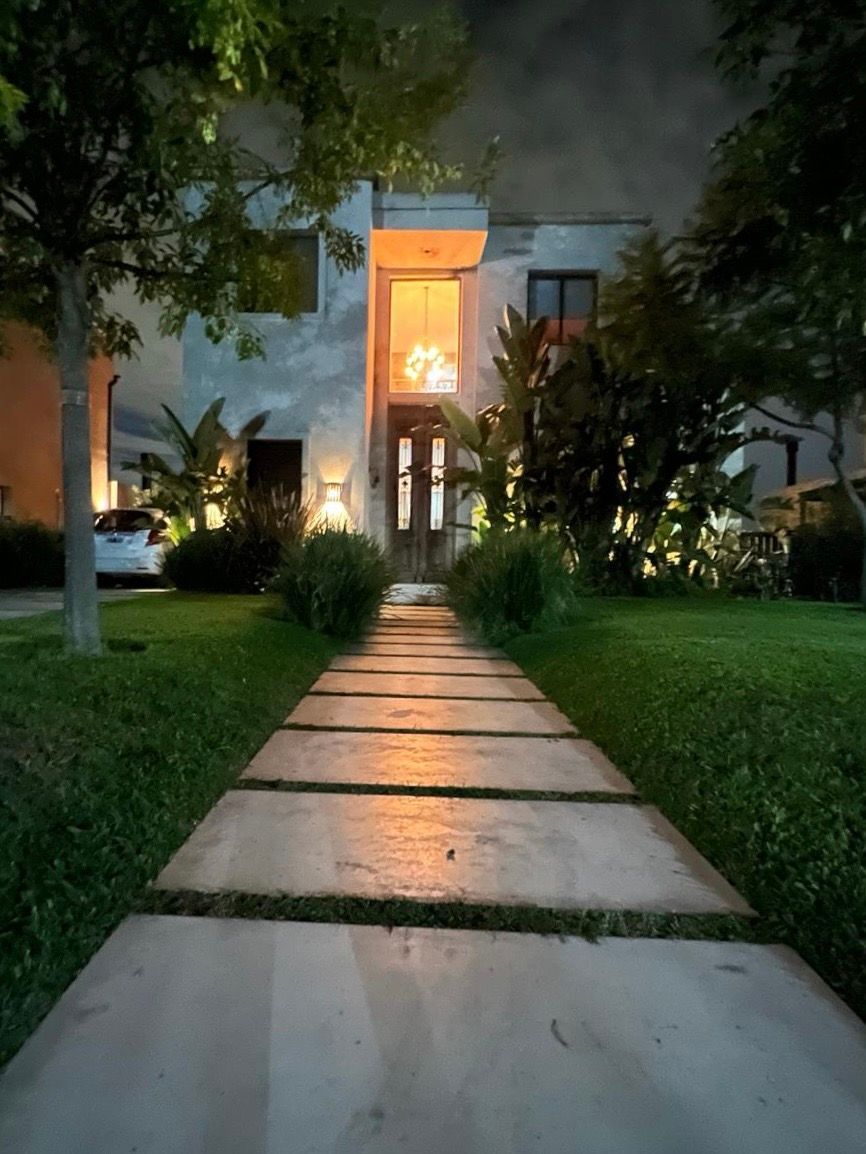 Sendero de piedra que conduce a una casa con una puerta iluminada, flanqueada por hierba verde y árboles al atardecer.