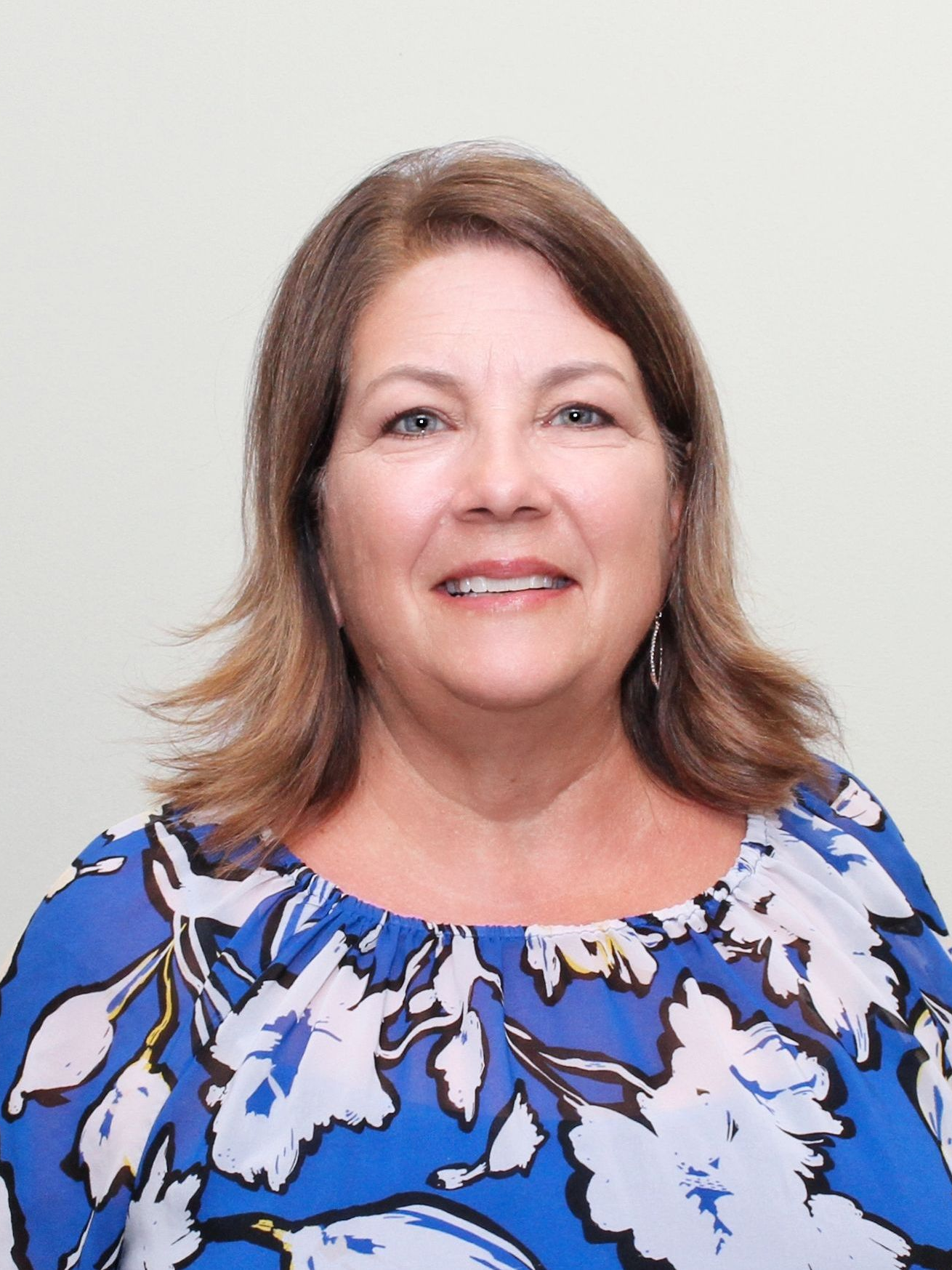 Woman with brown hair smiles, wearing a blue floral top, standing in front of a neutral wall.