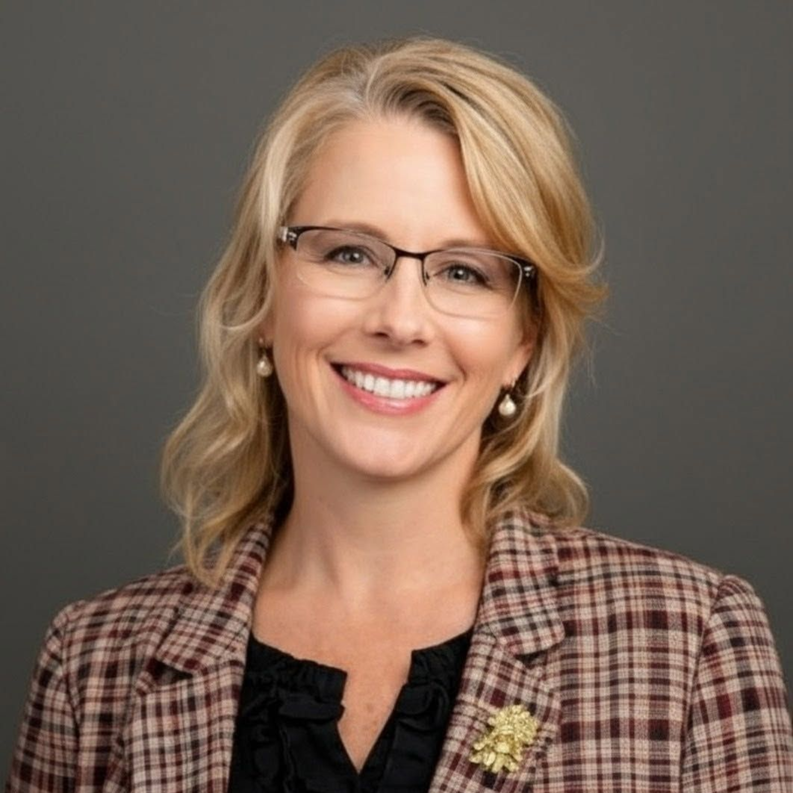 Woman with blonde curly hair, glasses, smiling, wearing a gray blazer and red top.