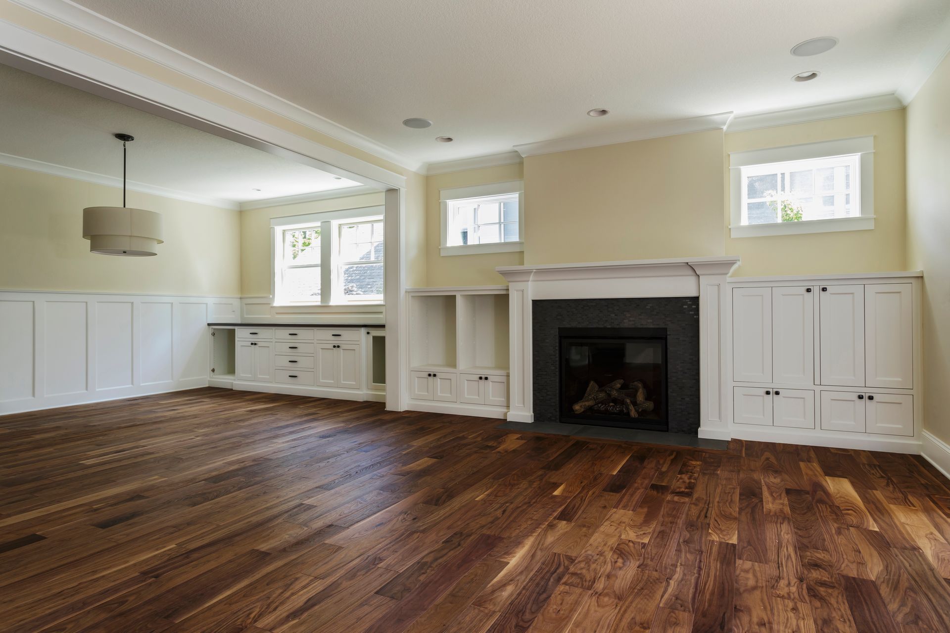 Empty living room with wood floors, fireplace, white built-ins, and two windows.