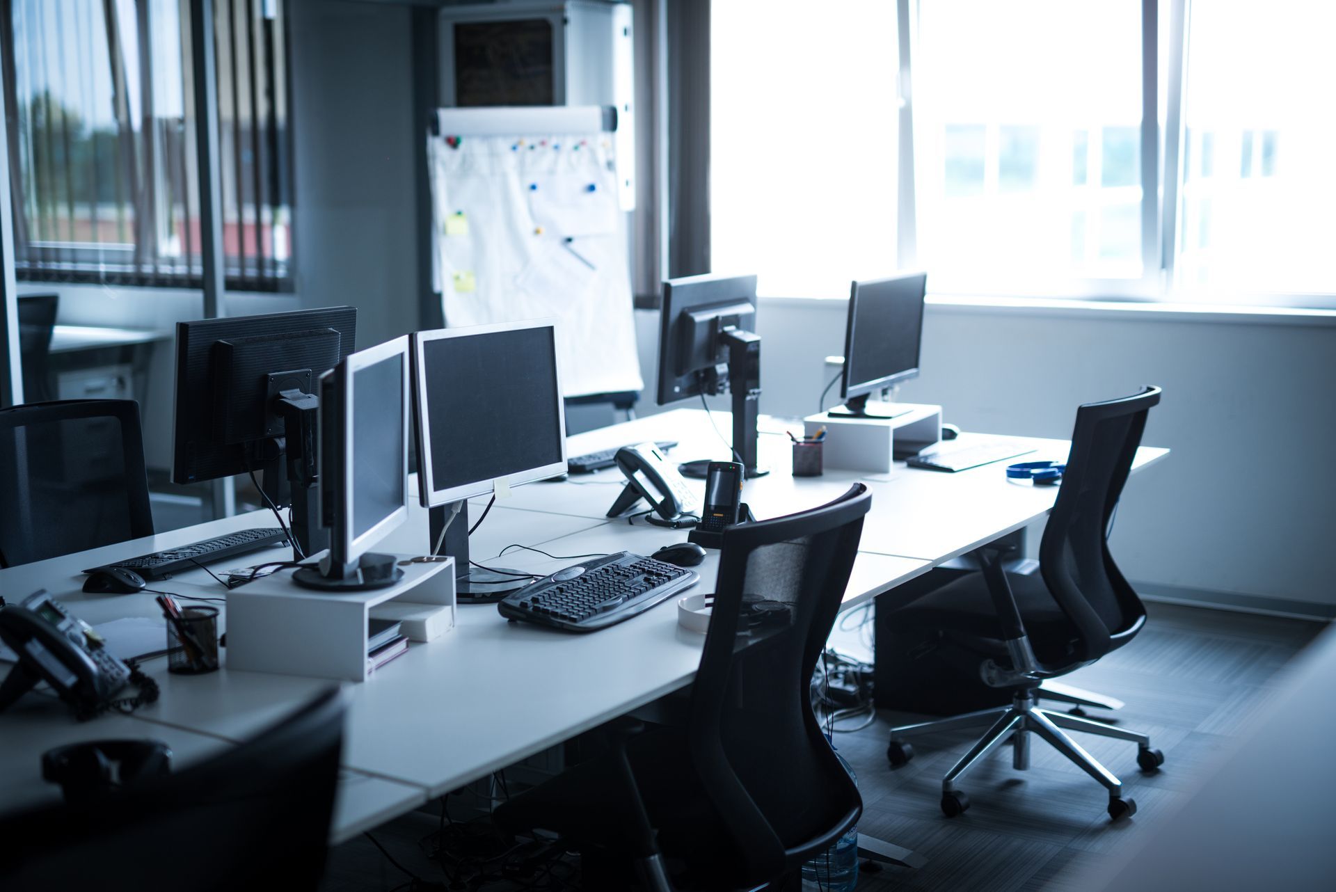 Office desk with multiple computer monitors, keyboards, and chairs near a window.