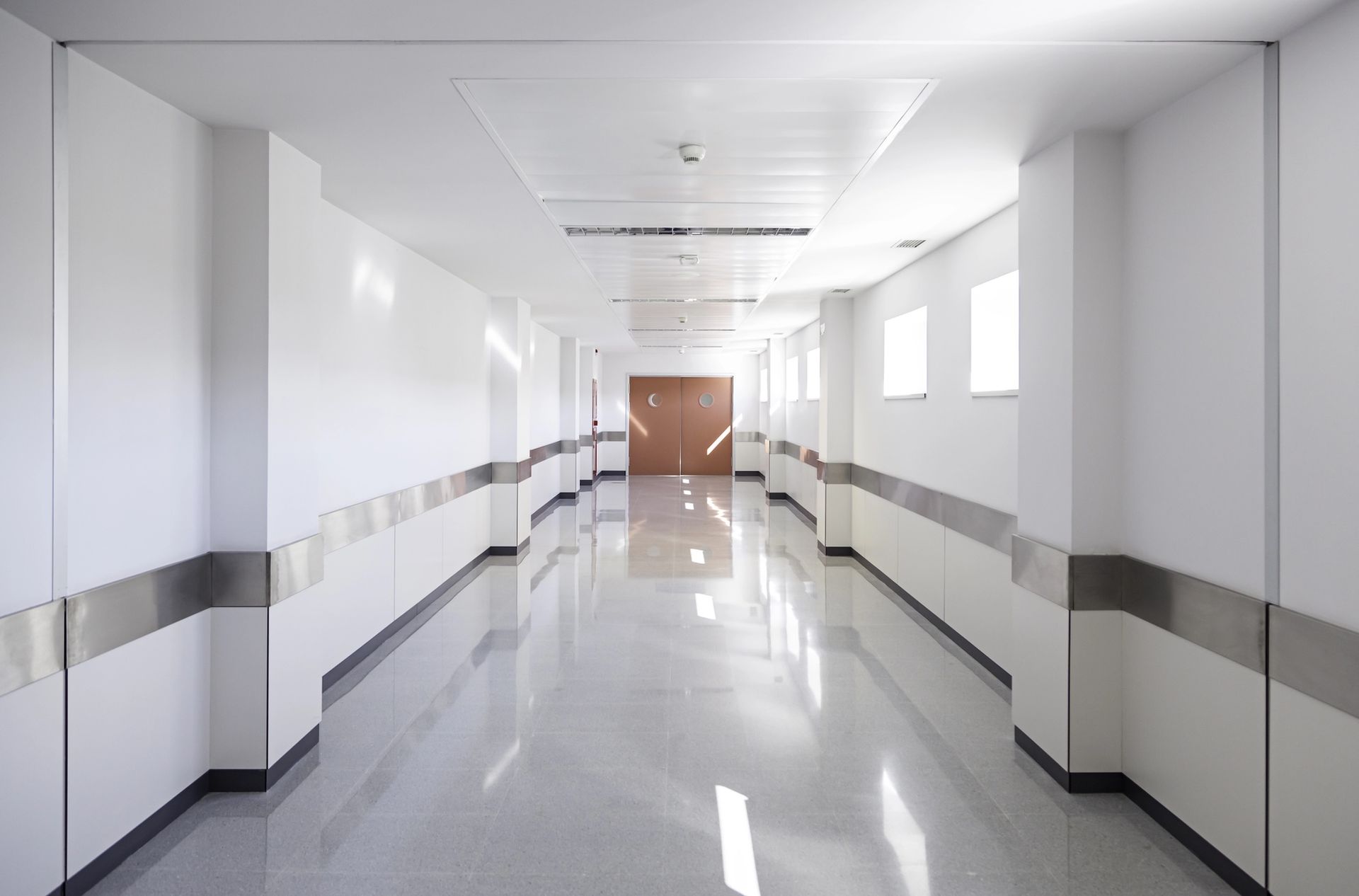 Bright, empty hospital hallway with white walls, reflective floor, and a closed wooden door at the end.
