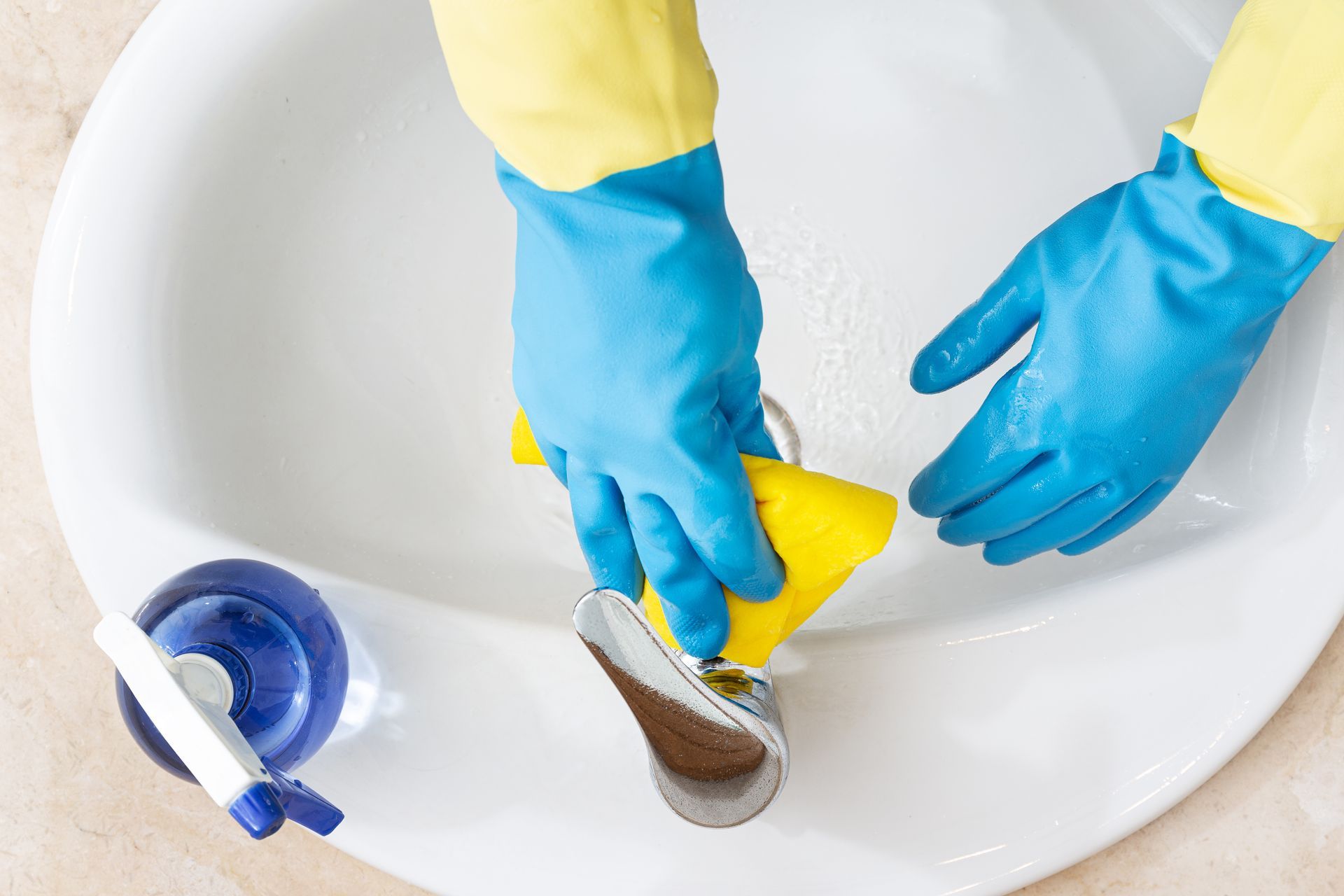 Hands in blue gloves cleaning a white sink faucet with a yellow sponge.