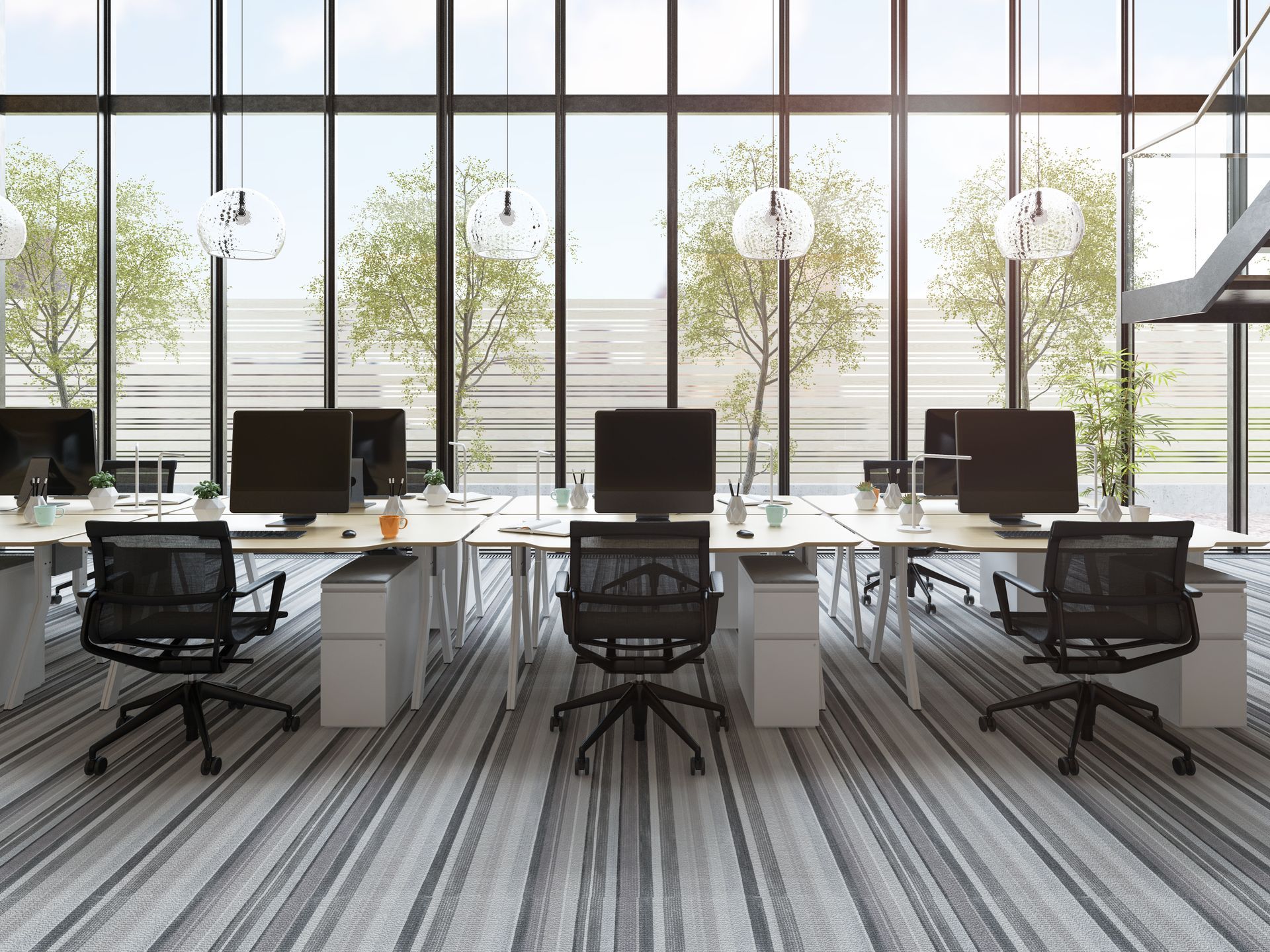 Office desks with computers, black chairs, large windows overlooking trees.