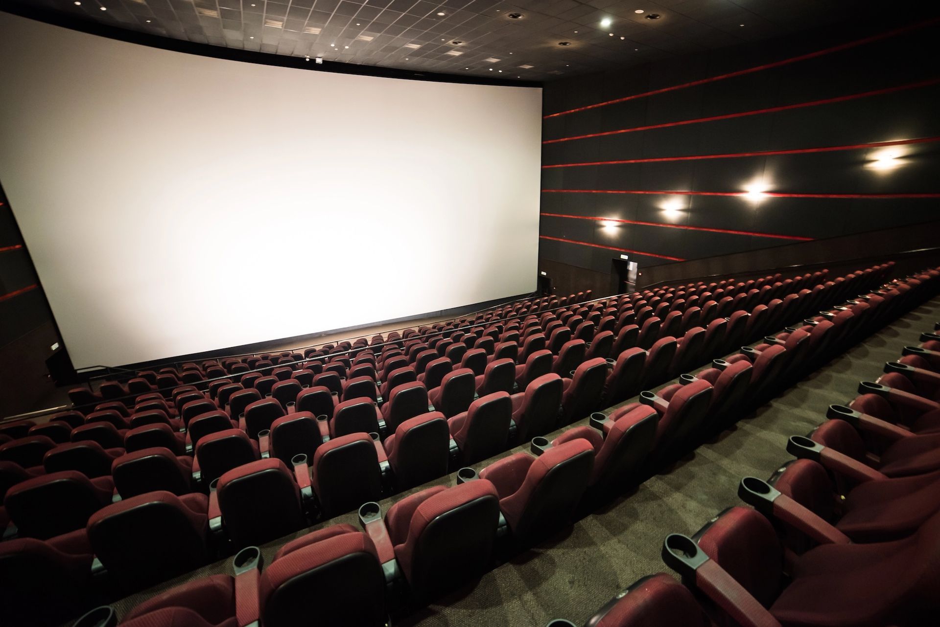 Empty movie theater with red seating and large white screen.
