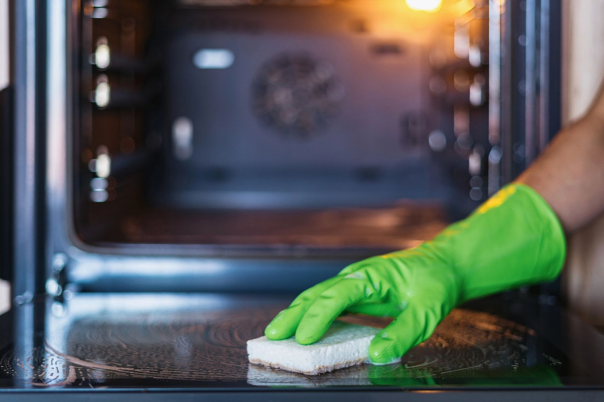 Hand in green glove cleaning inside of oven with a sponge.
