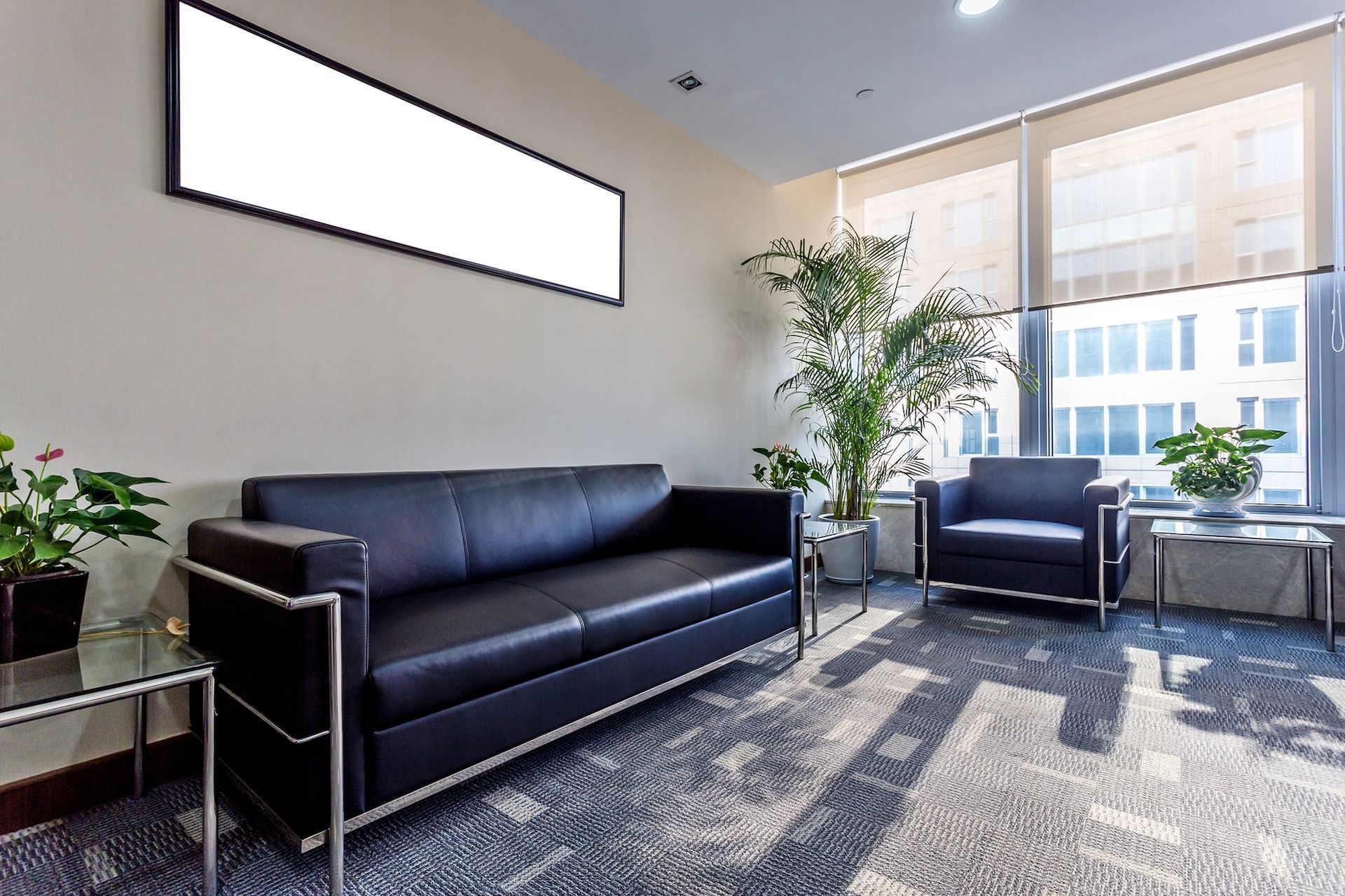 Waiting room with a black leather couch, armchair, plants, and a window. A blank sign is above the couch.
