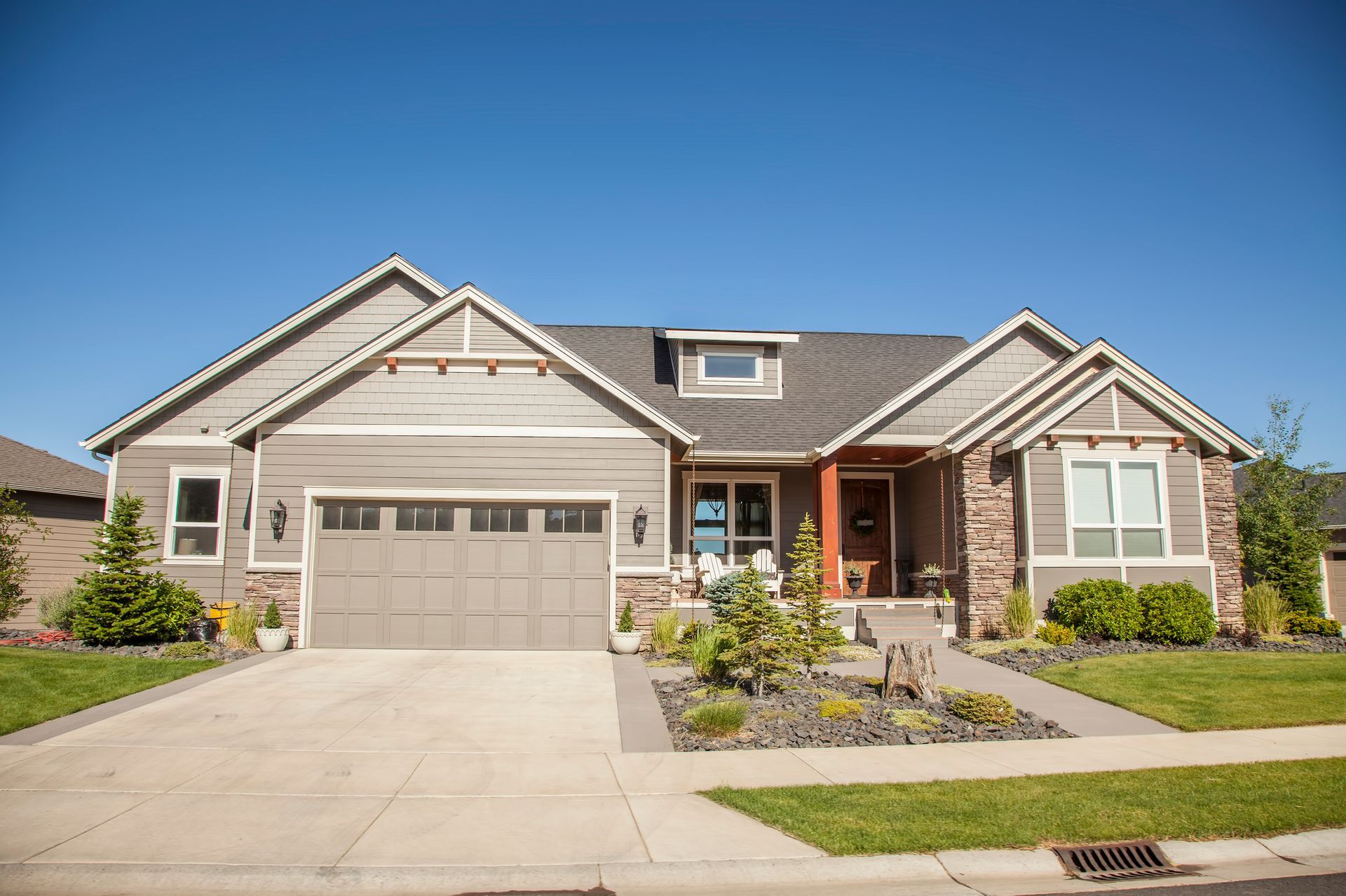 Gray house with a brown garage door, brick accents, and a landscaped front yard under a blue sky.