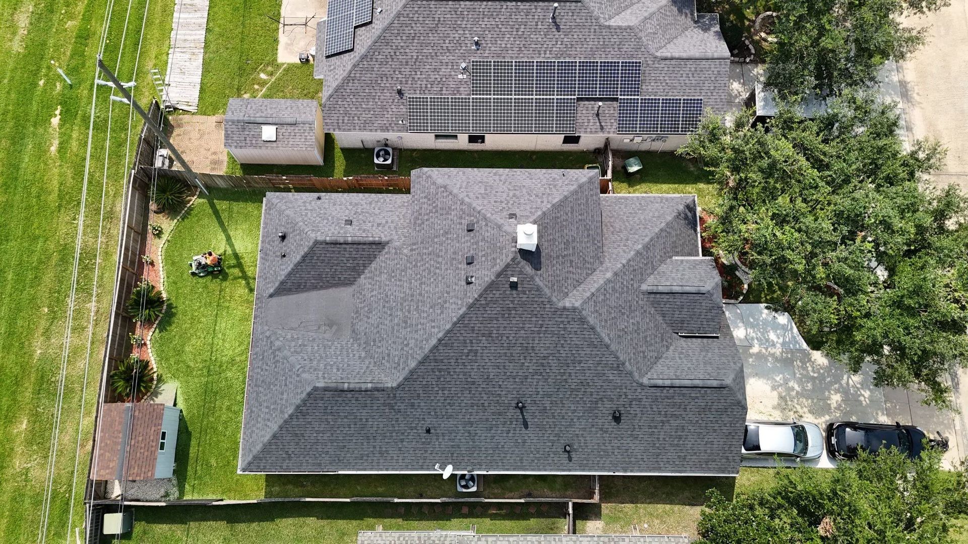 Aerial view of a house with a dark gray roof, surrounded by green grass, other houses, and trees.
