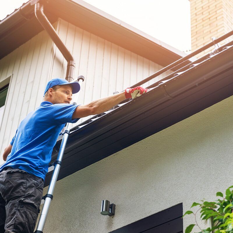 Man on a ladder cleaning gutters on a house with a blue shirt and cap, sunny day.