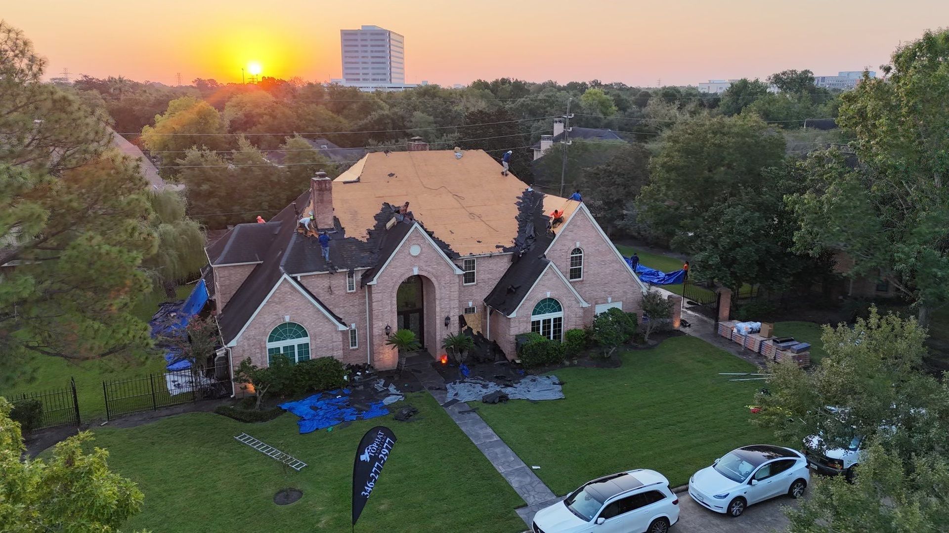 House undergoing roof repair at sunset; workers, blue tarps, cars, and trees in Houston, TX.