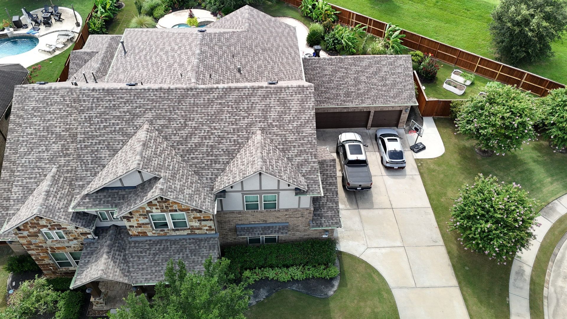 Aerial view of a large house with a new roof replacement, driveway, cars, and a pool in the backyard.