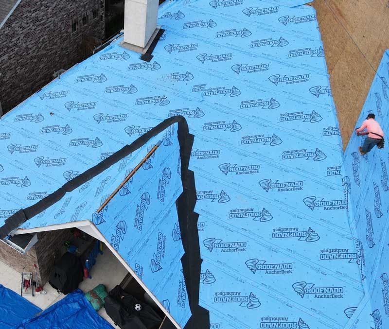 Blue roofing underlayment on a roof, with a worker kneeling at the right edge.