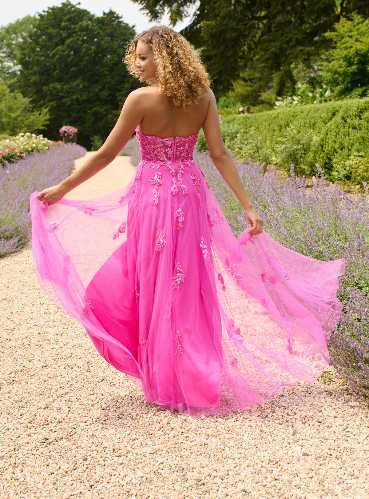 Woman in pink formal dress walking on path, holding up the skirt. Surrounded by lavender bushes.